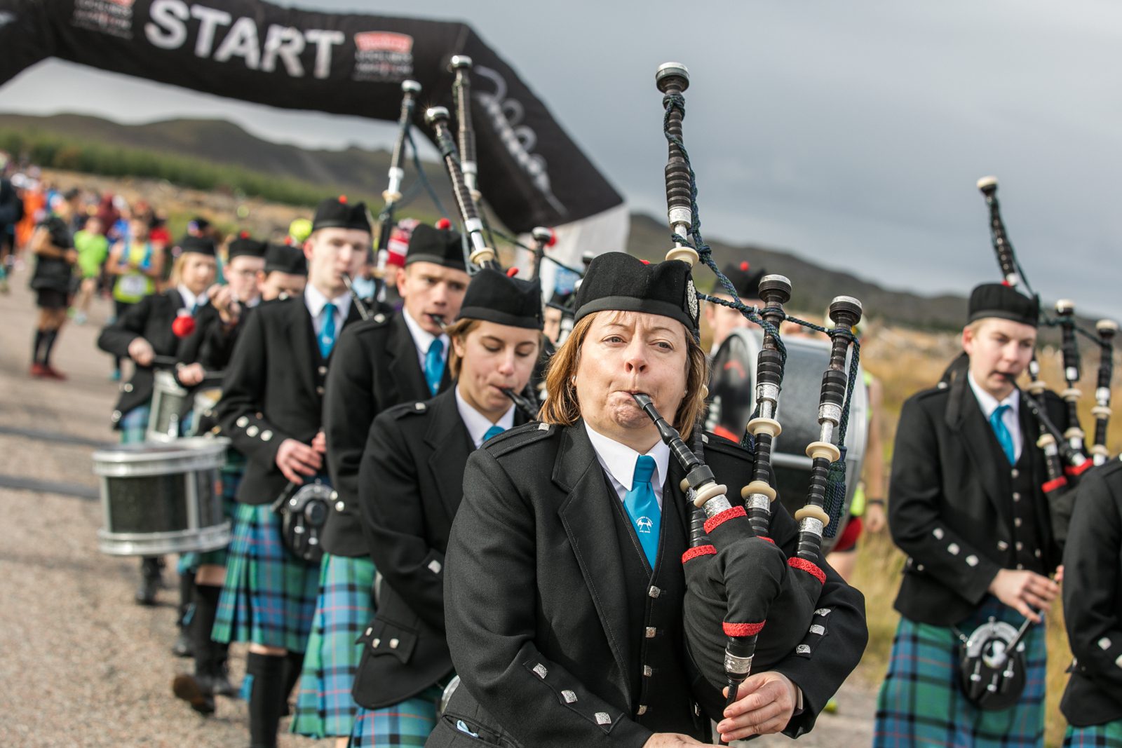 A group of people wearing traditional Scottish attire, including kilts and hats, are playing bagpipes and drums while marching. They are outdoors, and a "START" banner is visible in the background, suggesting they are part of an event or parade.