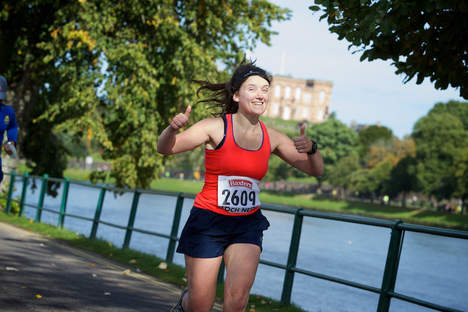 A smiling runner in a red tank top and black shorts is giving two thumbs up while running on a pathway beside a river. She is wearing a race number 2609 pinned to her shirt and appears to be enjoying the event. Trees and a building are visible in the background.