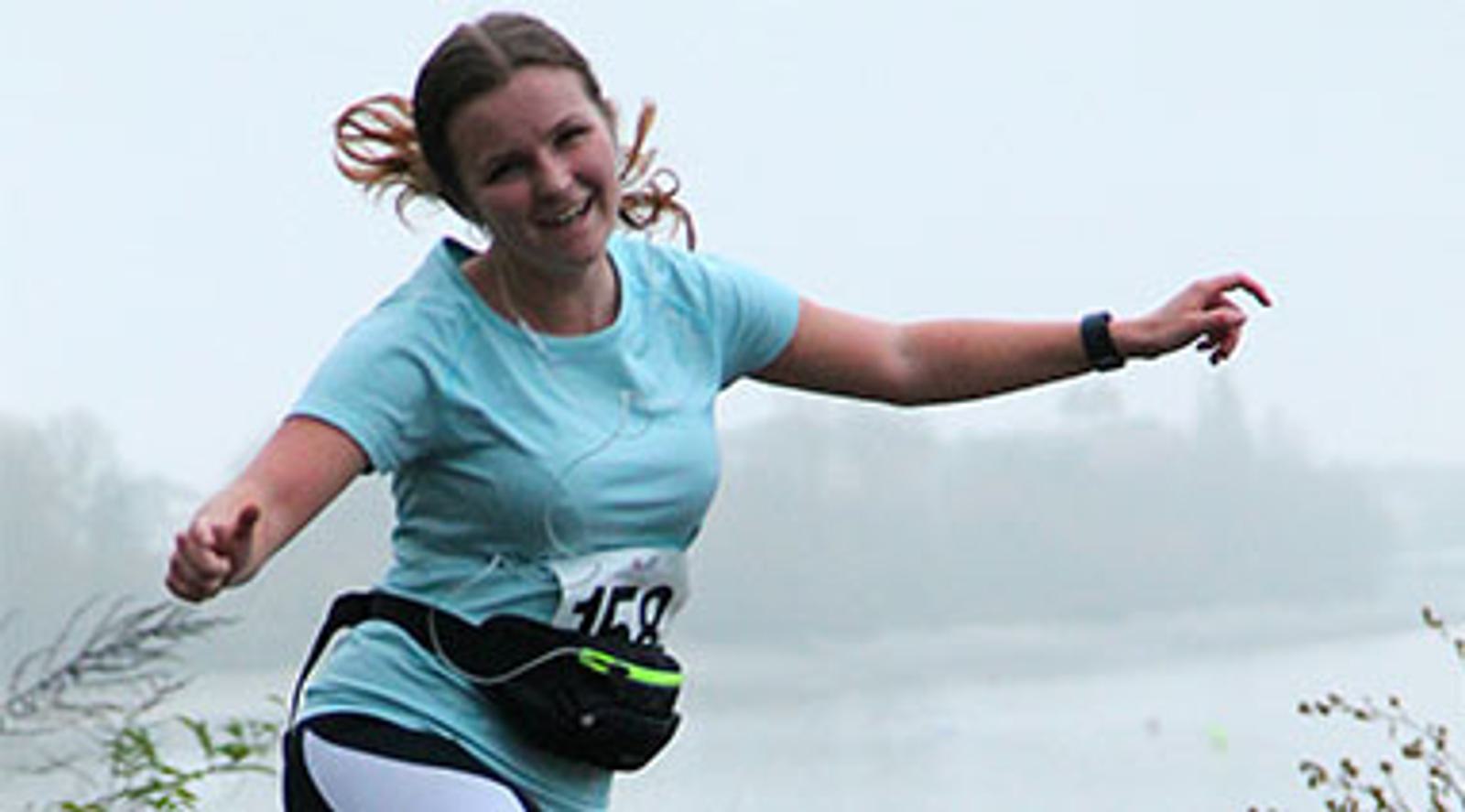 A woman in athletic wear running outdoors. She has a race number 148 on her shirt and a waist pouch. The background is a misty landscape with water and trees. She is smiling and appears to be mid-stride, with her arms outstretched.