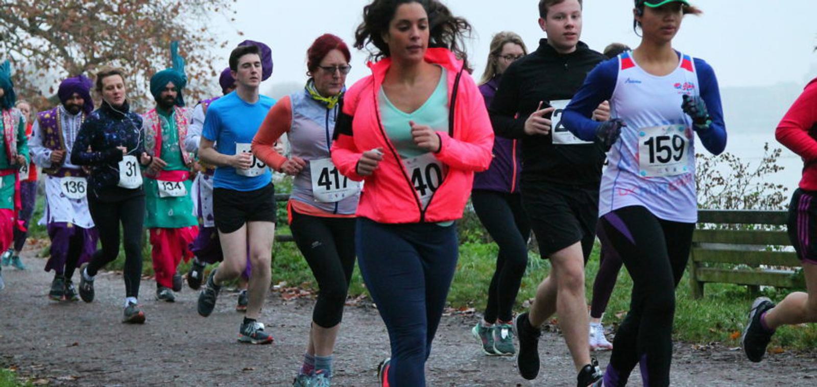 A group of runners, including men and women, participate in an outdoor race on a gravel path near a body of water. The participants wear athletic gear, including jackets, leggings, and running shoes. Trees with autumn leaves are visible along the path.