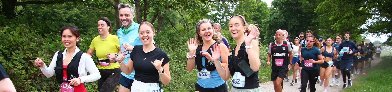 A group of runners, smiling and waving, participate in an outdoor race along a tree-lined path. They wear race bibs and athletic gear, with greenery and other runners visible in the background.