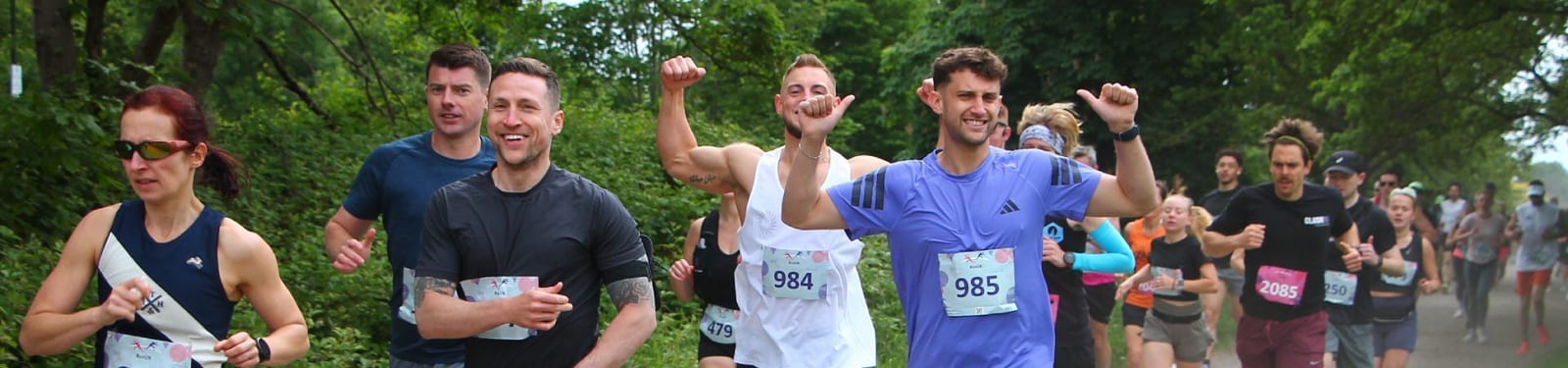 A group of runners, some smiling and raising their arms in celebration, participate in a race on a tree-lined path. Each person wears a numbered bib, and the atmosphere is energetic and cheerful.