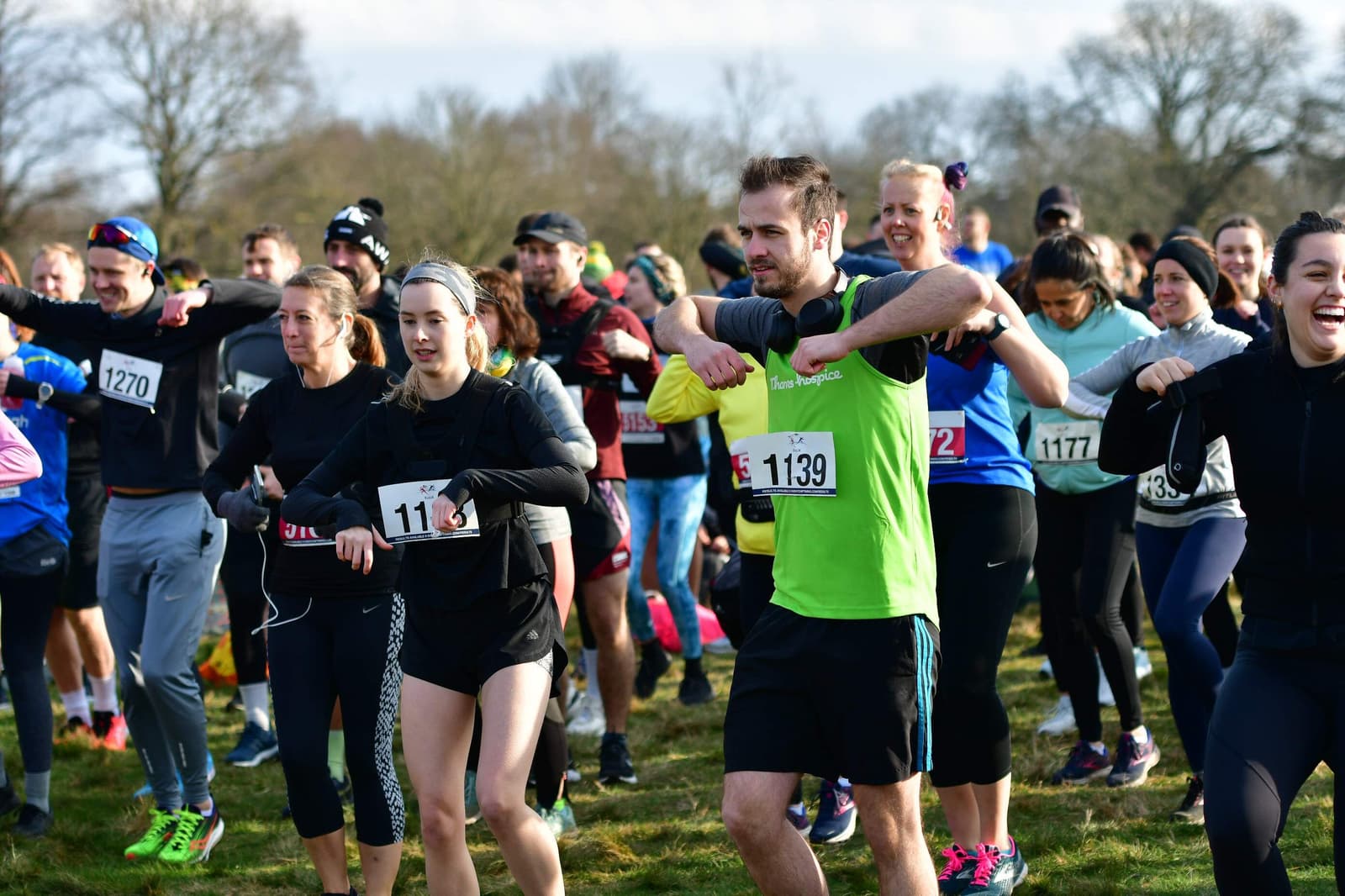 A large group of runners warming up for a race in an outdoor setting. They are wearing various athletic outfits and numbered bibs. The weather appears cool, with some participants in jackets and others in short sleeves. Trees are visible in the background.