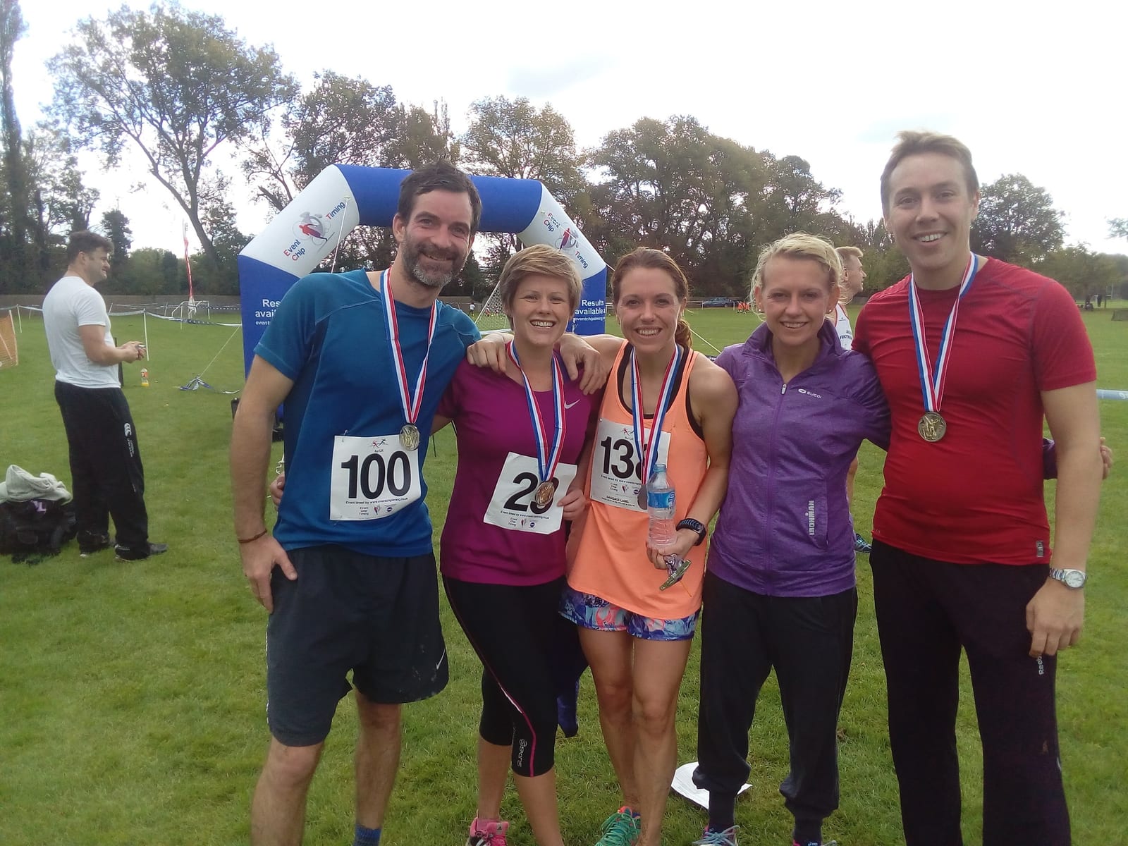 Five smiling people wearing race bibs and medals stand together on a grassy field, with an inflatable arch and trees in the background. They are dressed in athletic wear, indicating participation in a running event.