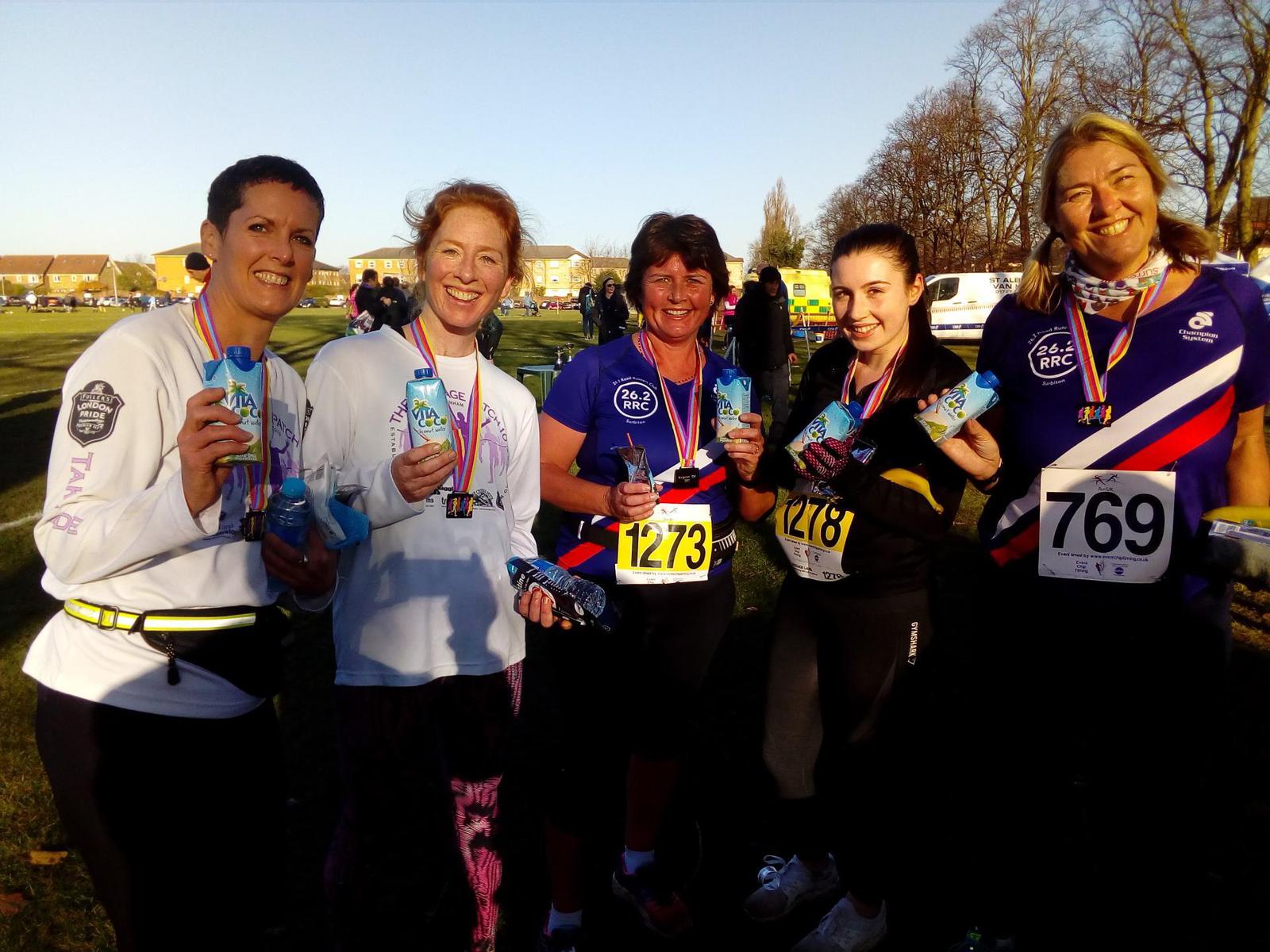 A group of five people, wearing race bibs and medals, smiles at the camera holding blue drinks. They are standing outdoors on a sunny day with trees and buildings in the background. The participants wear different athletic outfits, suggesting they have completed a race.