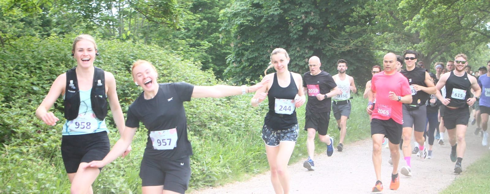 A group of runners, some smiling and reaching out playfully, participate in a race along a tree-lined trail. The atmosphere is lively and cheerful. Bib numbers are visible on their shirts.