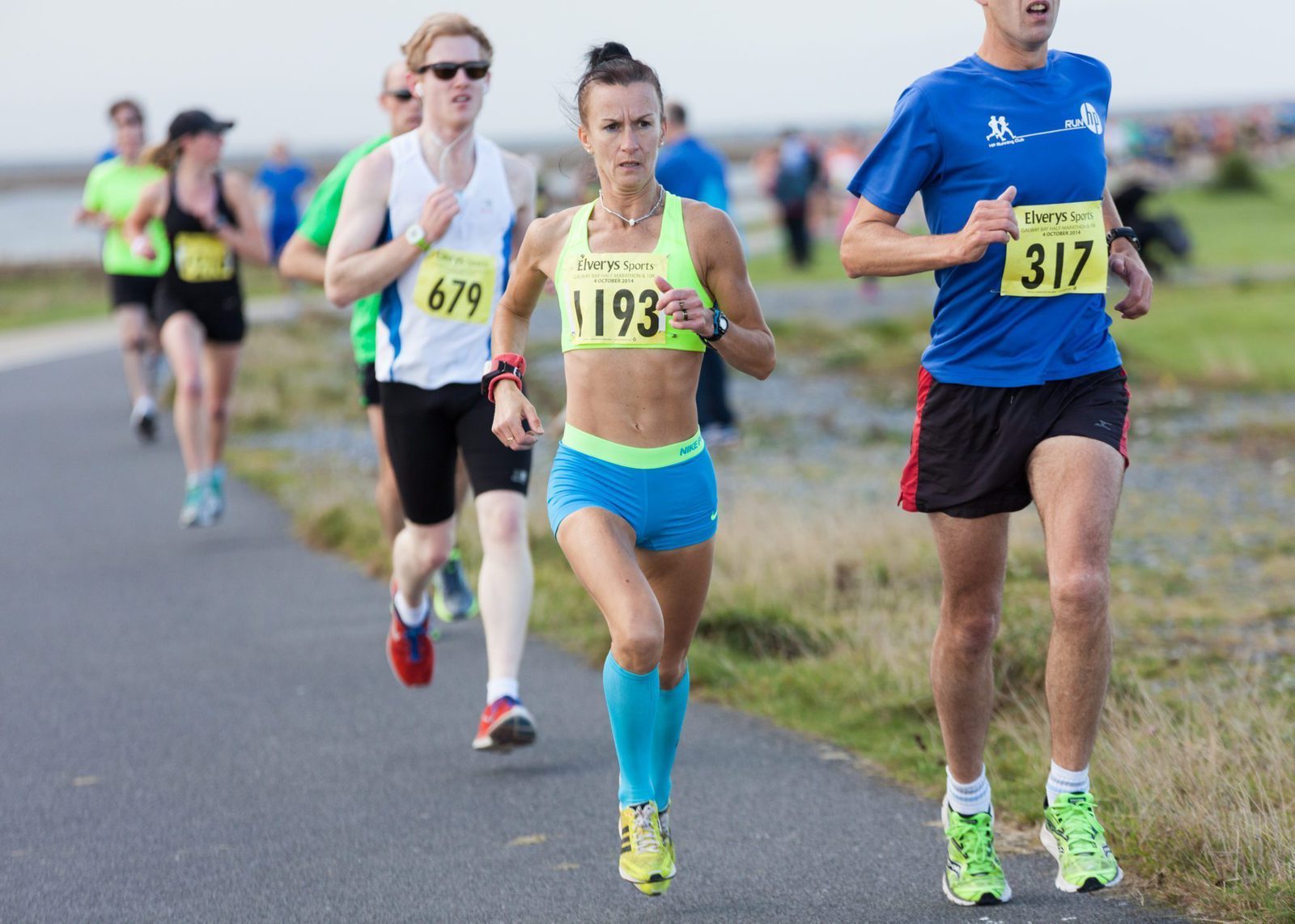 A group of runners is participating in a race on a paved path near the coastline. The runner in the foreground, wearing a yellow sports bra and blue shorts, appears focused. Other runners, both male and female, are visible in the background.