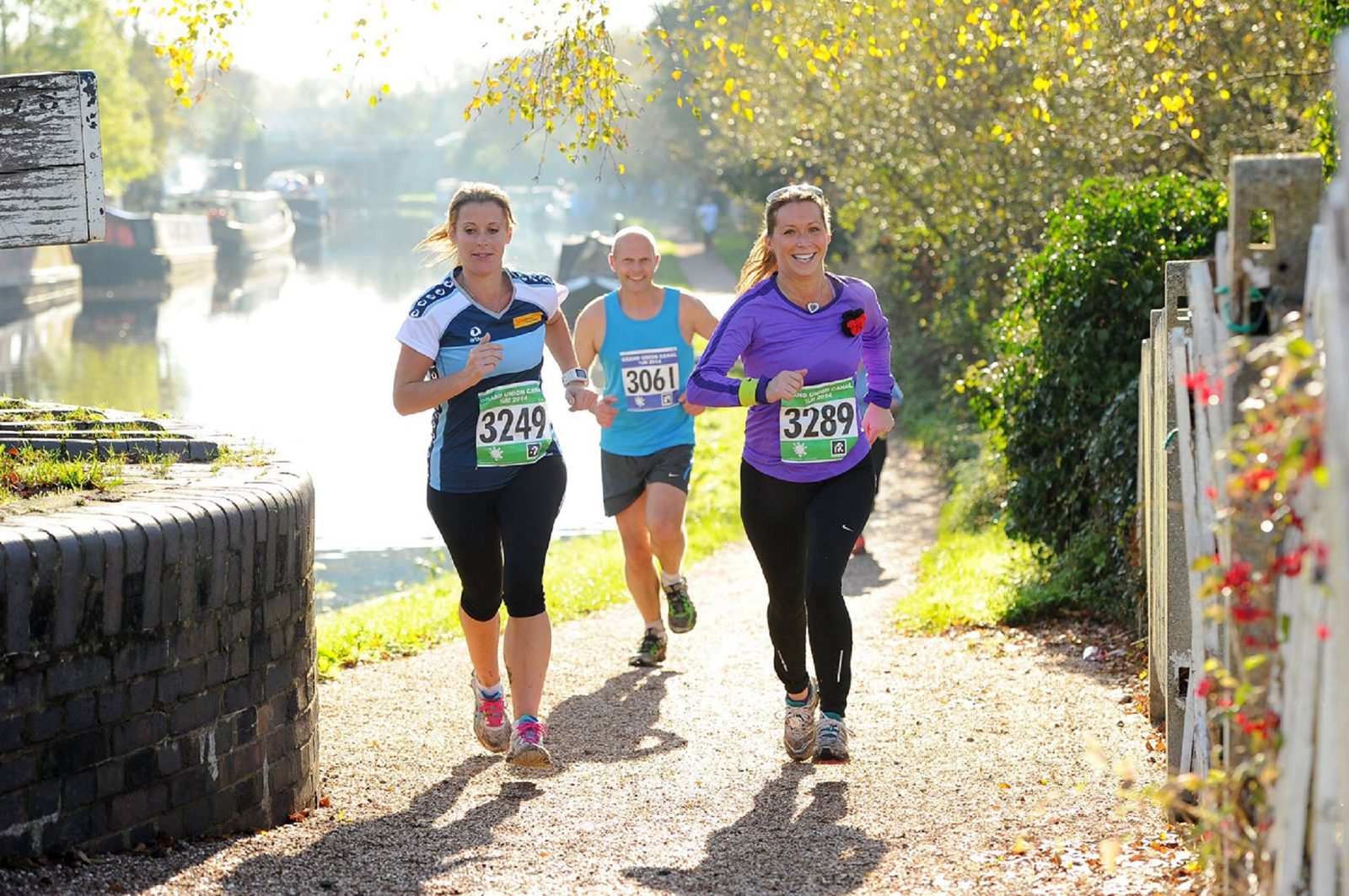 Three runners, all wearing numbered bibs, jog on a gravel path beside a canal under sunny skies. Two women in front, one in a blue shirt and the other in a purple shirt, smile towards the camera. A bald man in a blue shirt follows closely behind. Trees and a bridge are visible.