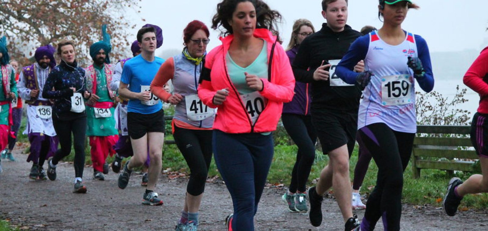 A group of people running in a race on a tree-lined path. The participants are wearing various sports clothing, some with numbers pinned to their shirts. The scene suggests an outdoor event, with leaves scattered on the ground and a slightly overcast sky.