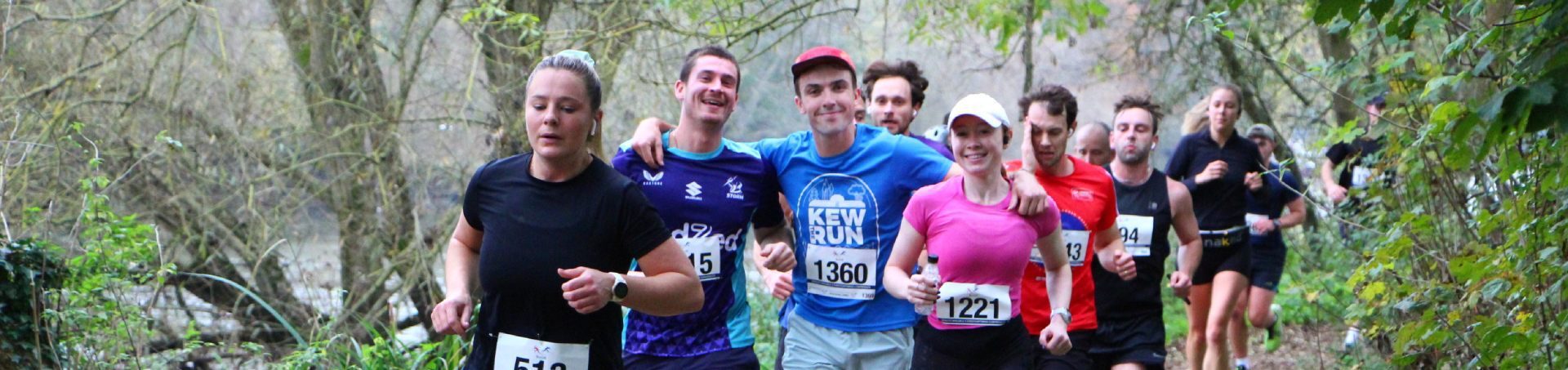A group of people run outdoors on a trail, smiling and wearing numbered race bibs. Trees and greenery line the path, creating a natural, scenic background.