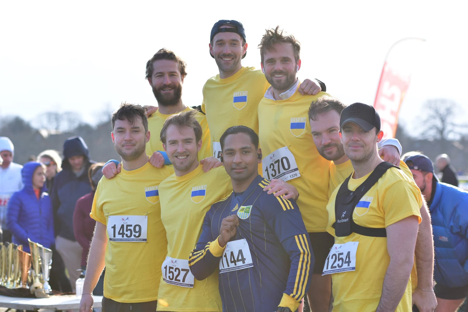 A group of eight men, wearing matching yellow sports shirts and participant bibs, pose together for a photo at an outdoor event. Some of the men are standing while others are kneeling in front. There are tents and people in the blurred background.