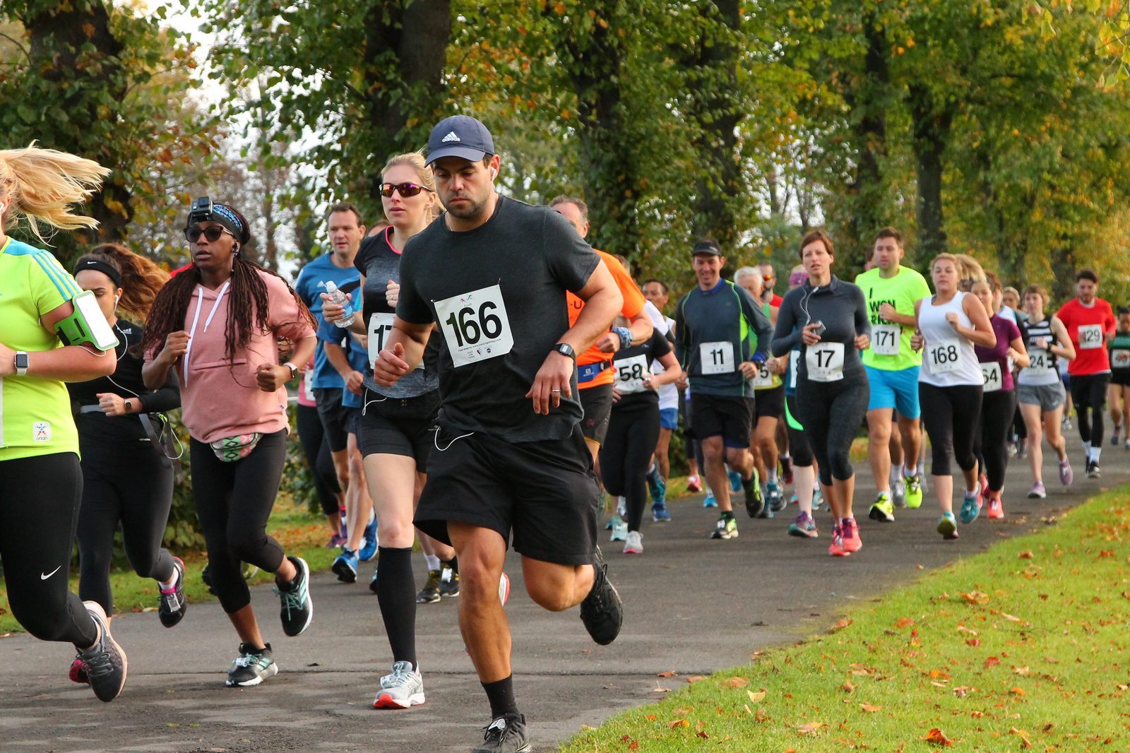 A diverse group of people running a race on a tree-lined path. They are all wearing numbered bibs and various athletic gear. Some participants look focused on their run, while others appear more relaxed. The weather is clear, with fallen leaves scattered on the ground.