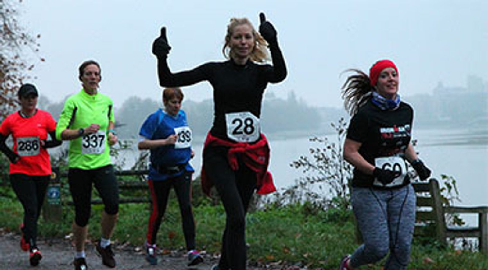 A group of runners participate in a race along a riverbank. One runner wearing a black outfit and number 28 smiles and gives thumbs up. Others, dressed in colorful athletic gear, run nearby. The setting is overcast with a calm river in the background.