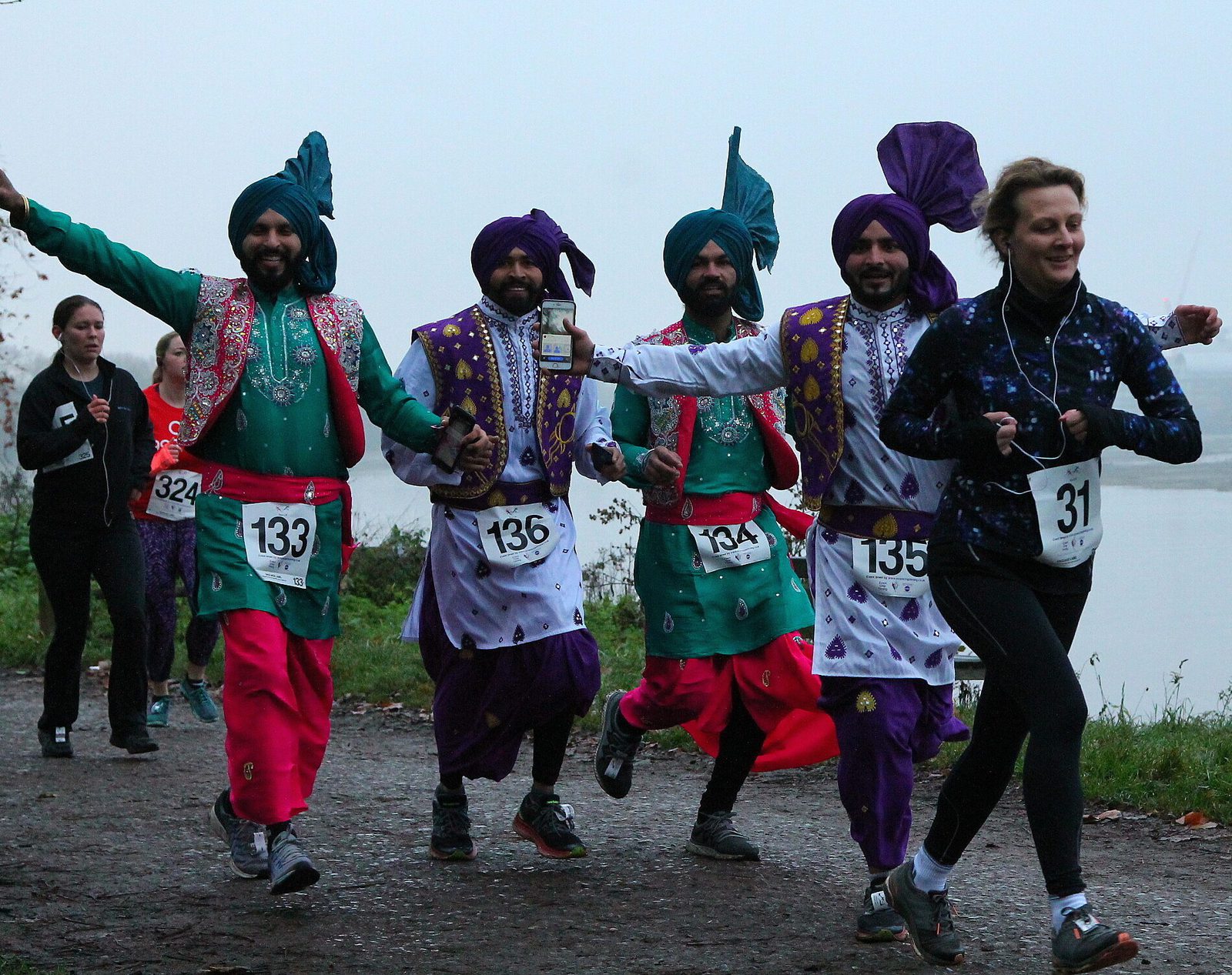 A group of four men dressed in vibrant traditional costumes joyfully run alongside a female runner on a foggy path beside a river. The men wear green and purple outfits with turbans, and the woman wears a black running outfit.
