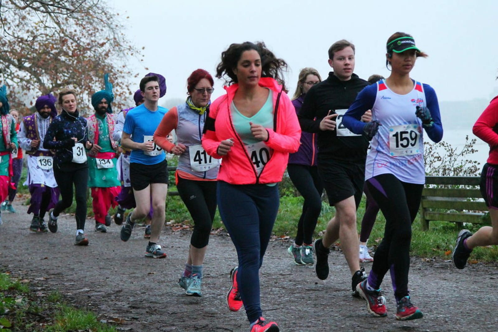 A group of runners participating in a race, with some wearing numbered bibs. The path is flanked by trees and a body of water is visible in the background. The runners are dressed in colorful athletic wear, and it appears to be a cloudy day.