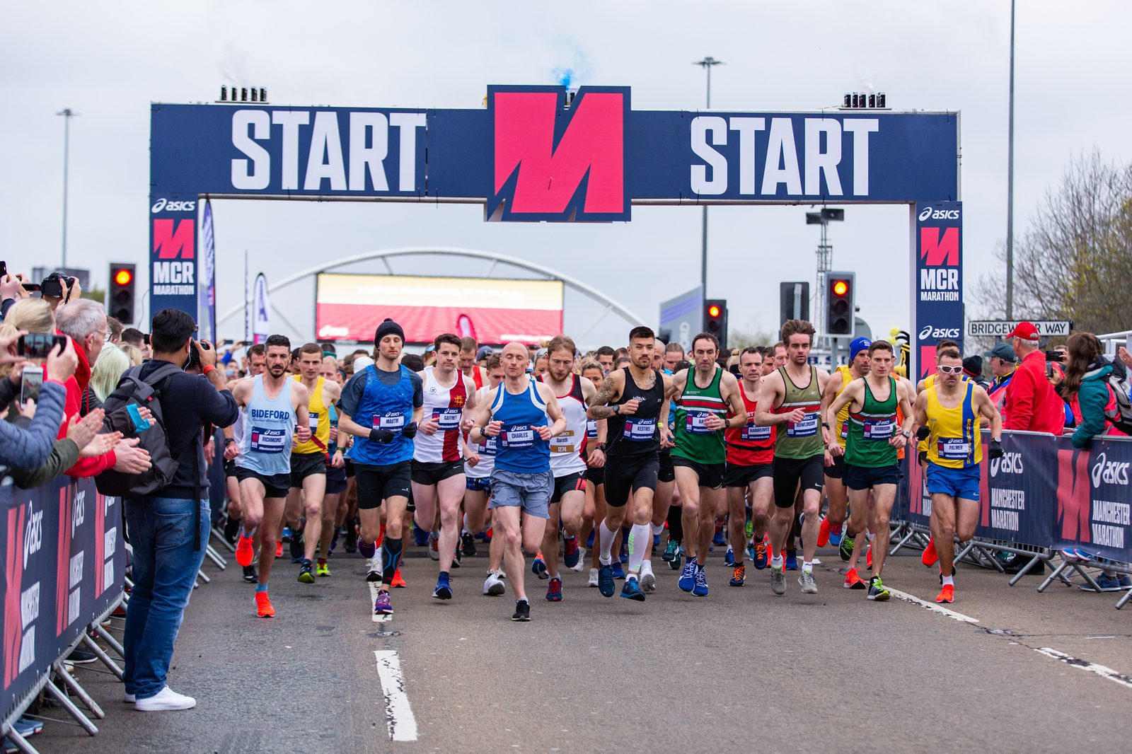A large group of runners at the starting line of a marathon race. The starting banner reads "START" with people in varied running attire poised to begin. Spectators with cameras and phones line the sides, and traffic lights and signage are visible in the background.