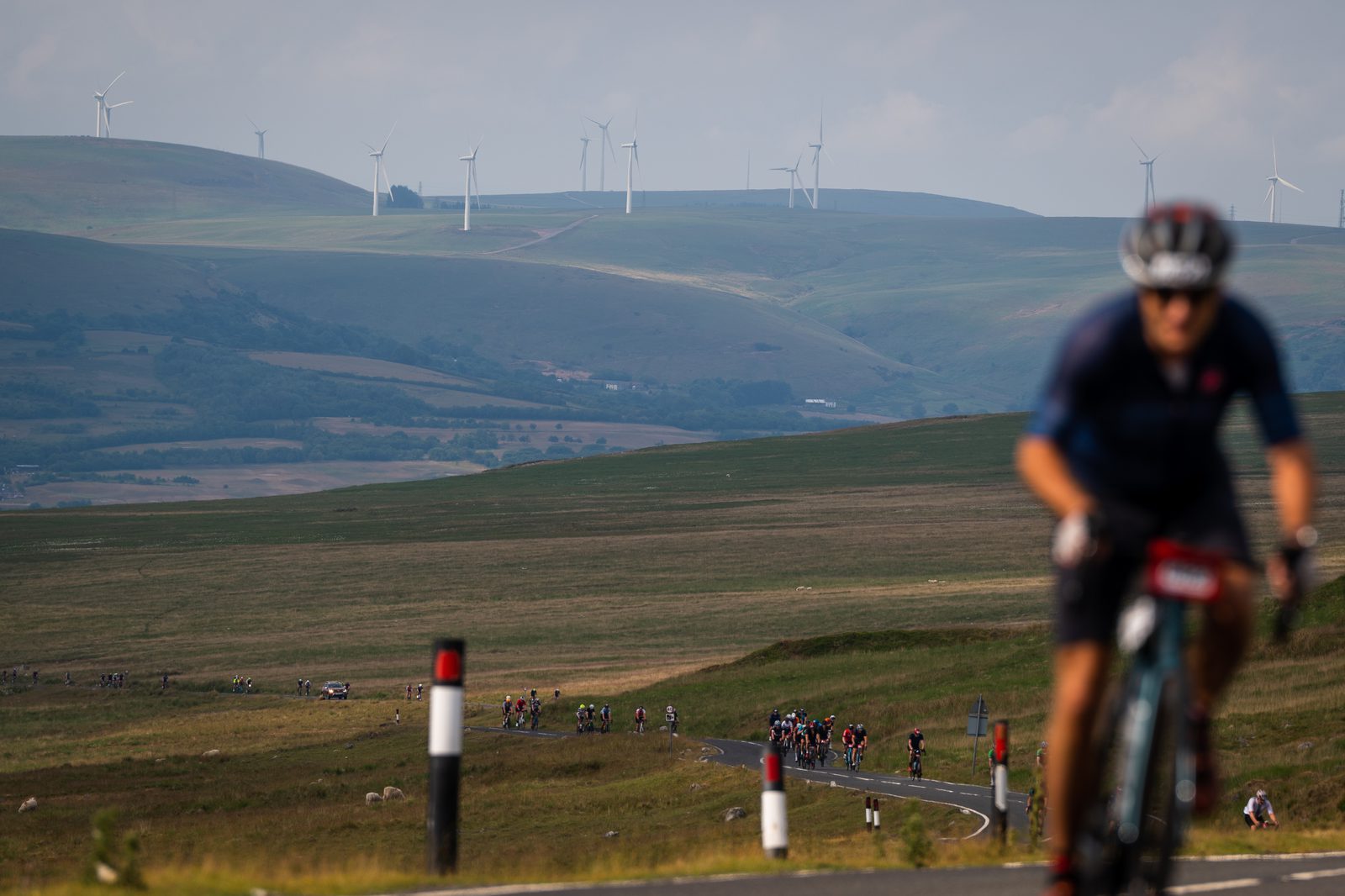A cyclist rides along a winding road in the foreground, with a group of cyclists in the distance. The landscape features green rolling hills topped with wind turbines under a partly cloudy sky. The scene captures the essence of an open countryside cycling event.