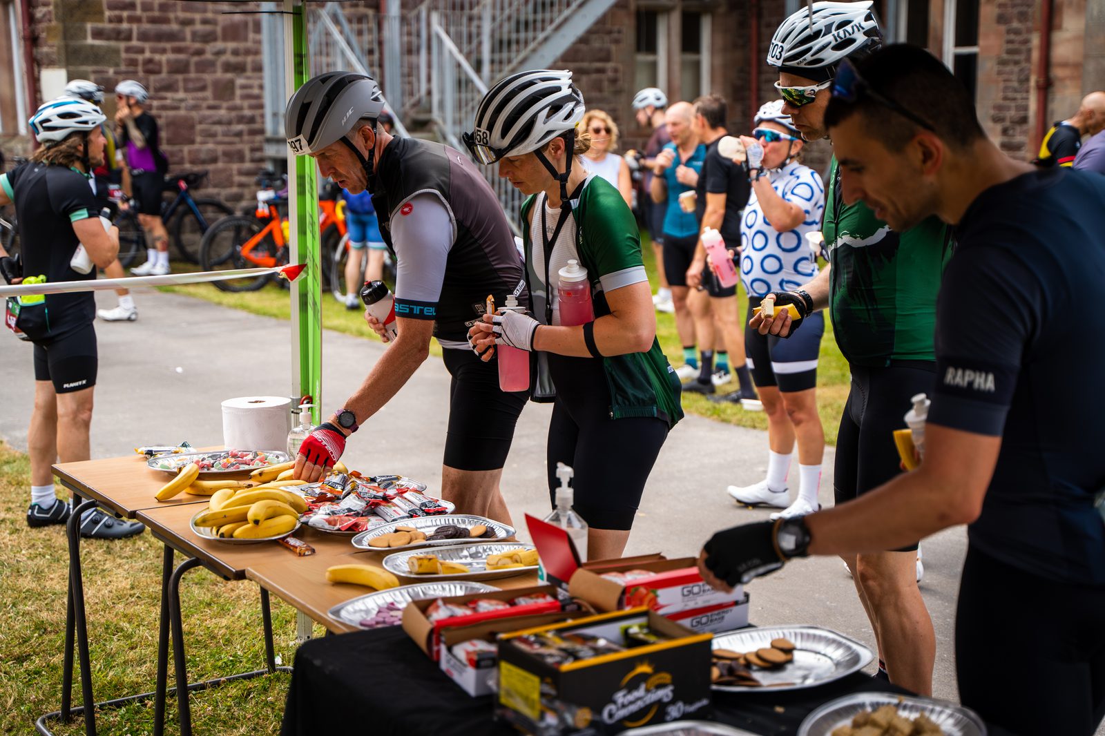 Cyclists in jerseys and helmets gather around a table stocked with food like bananas, nuts, and boxed pastries at an outdoor rest stop. Some are selecting food while others are chatting in the background near a building with a staircase.