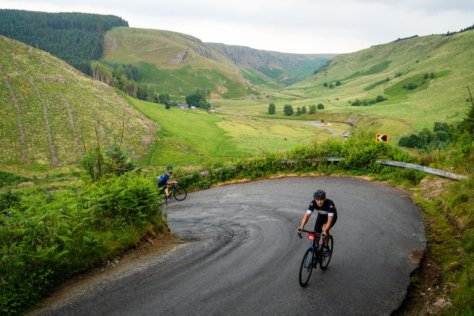 Two cyclists navigate a sharp curve on a hilly road surrounded by lush, green hills and valleys. One cyclist is in the foreground, wearing a black outfit and helmet, while the other is in the distance. The scenery includes trees and rolling countryside.