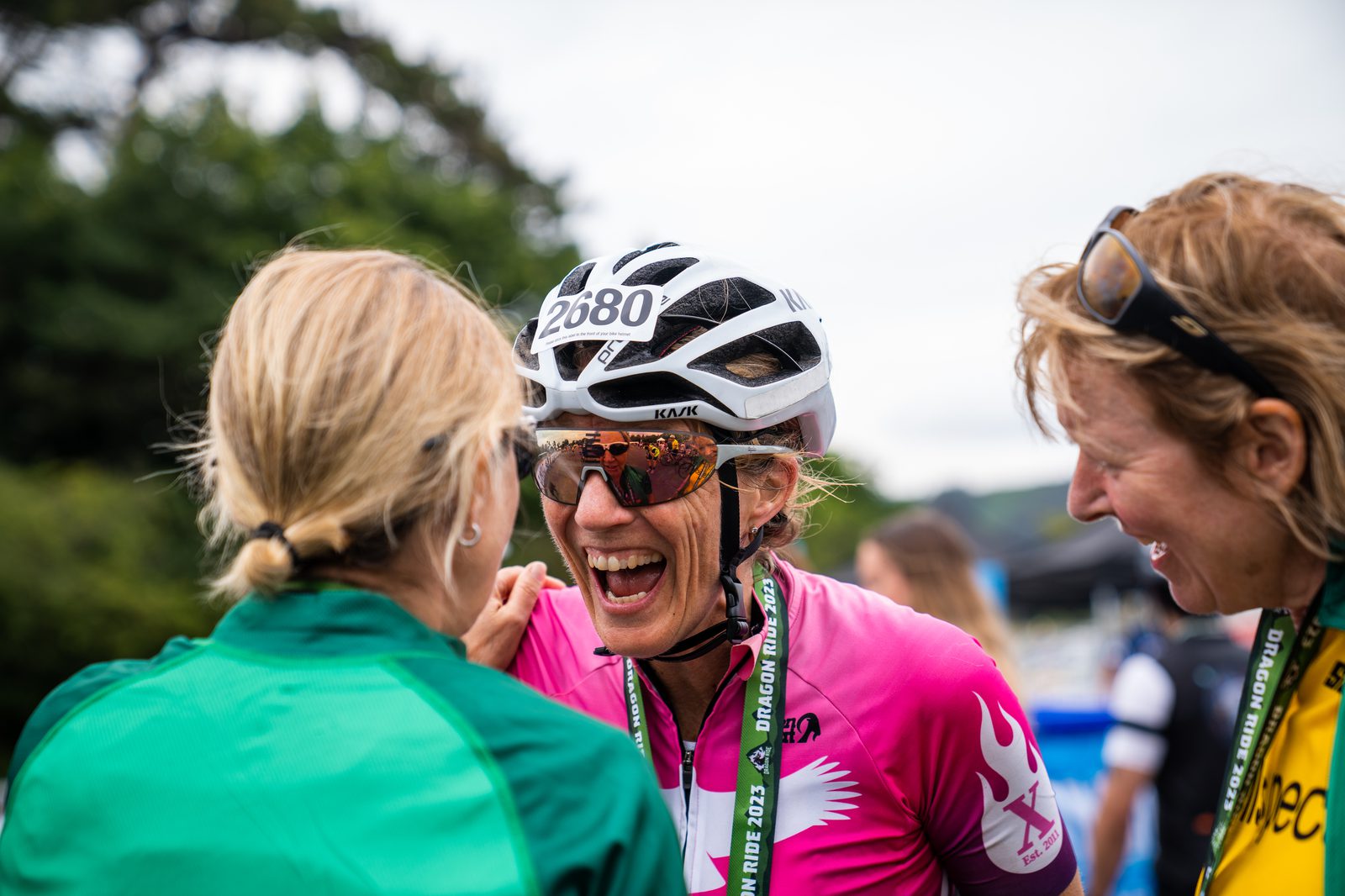 A cyclist wearing a white helmet, sunglasses, and a pink cycling jersey with the number 2680 on it is smiling and interacting with two other people. One of them wears a green top and the other has a yellow lanyard. The background has blurred greenery.