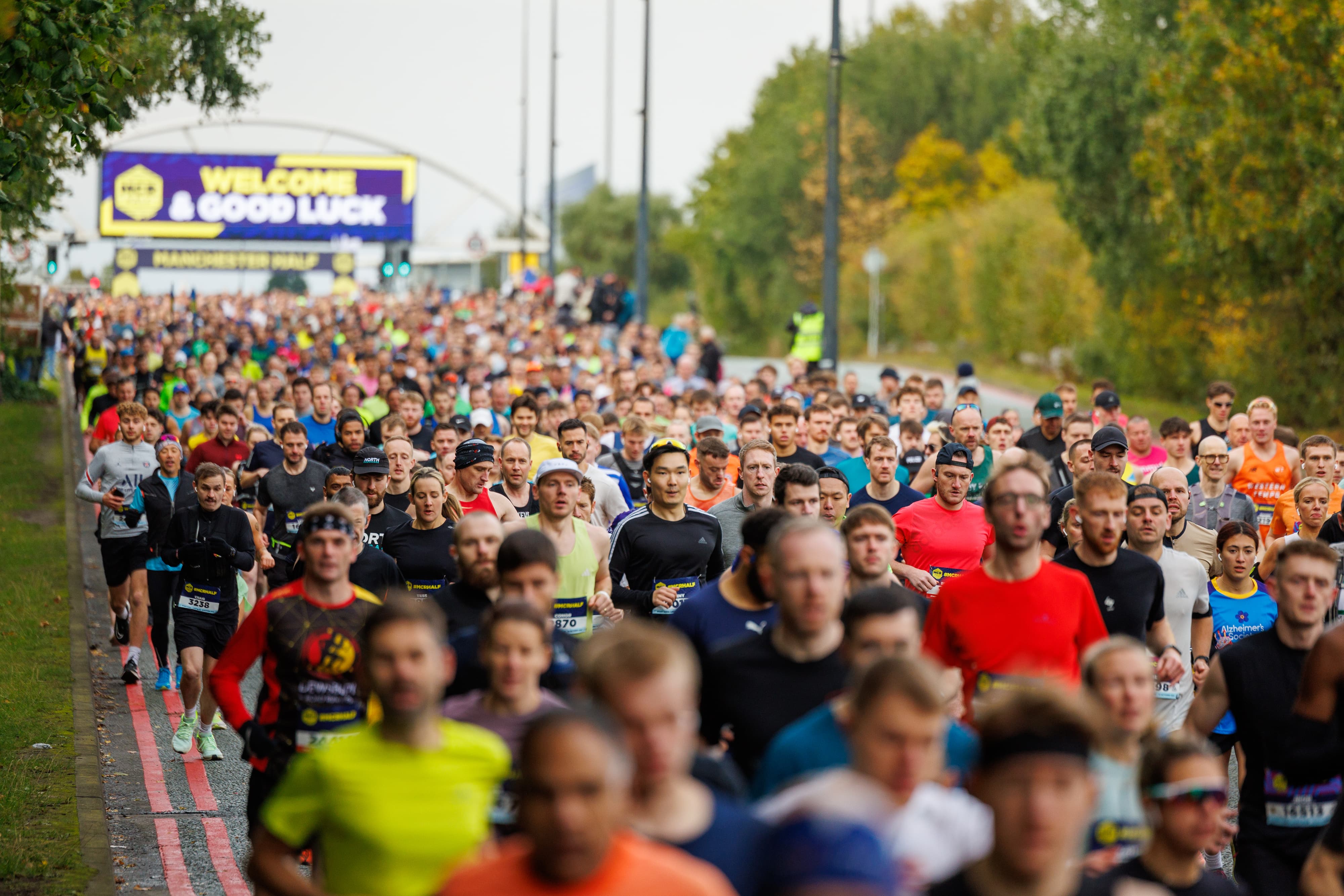 A large crowd of runners participates in a marathon on a road lined with trees. A prominent banner in the background reads "Welcome & Good Luck." The runners wear a variety of athletic gear in different colors.