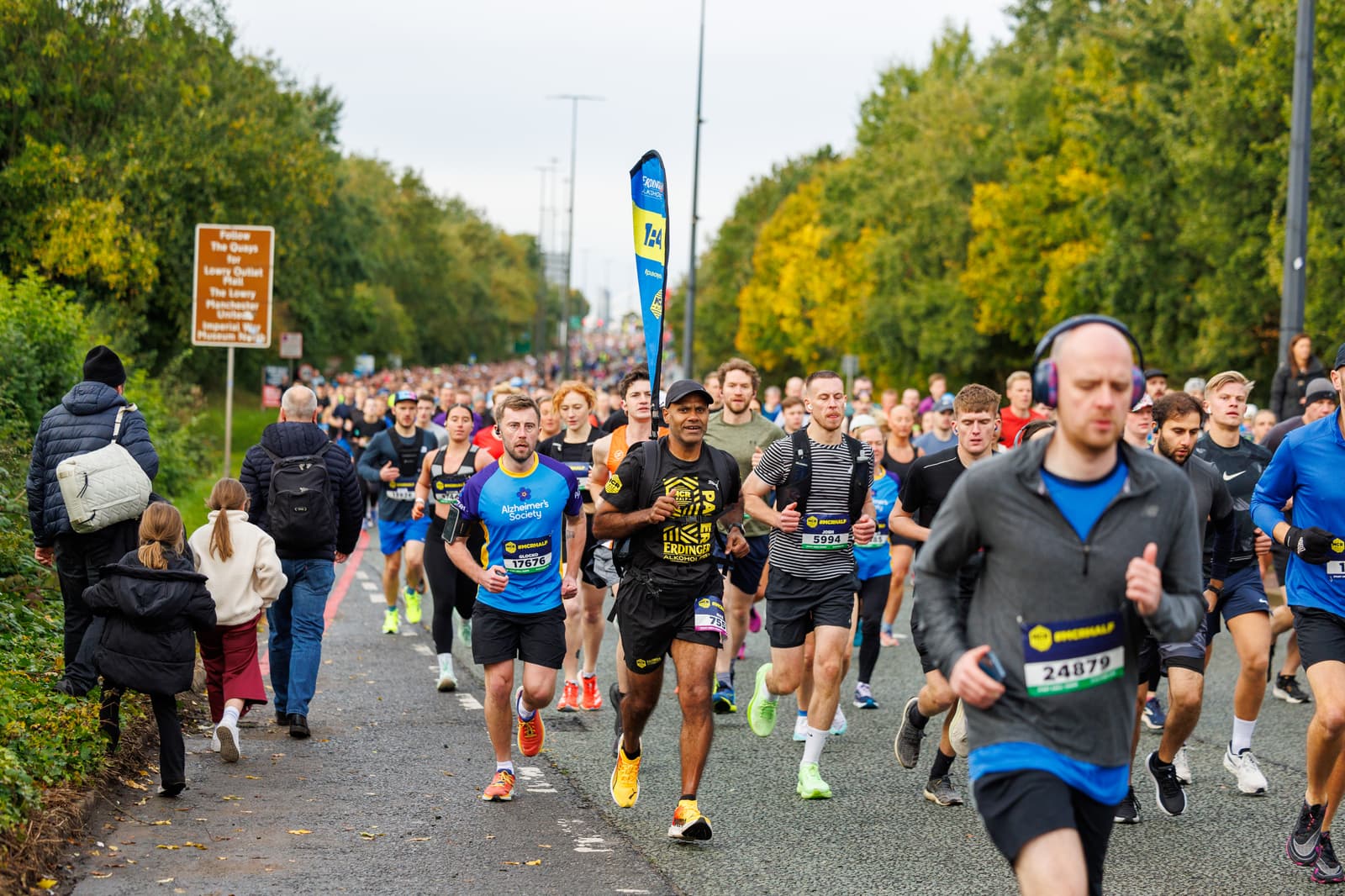 A large group of marathon runners of various ages run down a road lined with trees on a cloudy day. Some wear numbered bibs, and people are observing from the roadside. A man holding a flag leads the race.