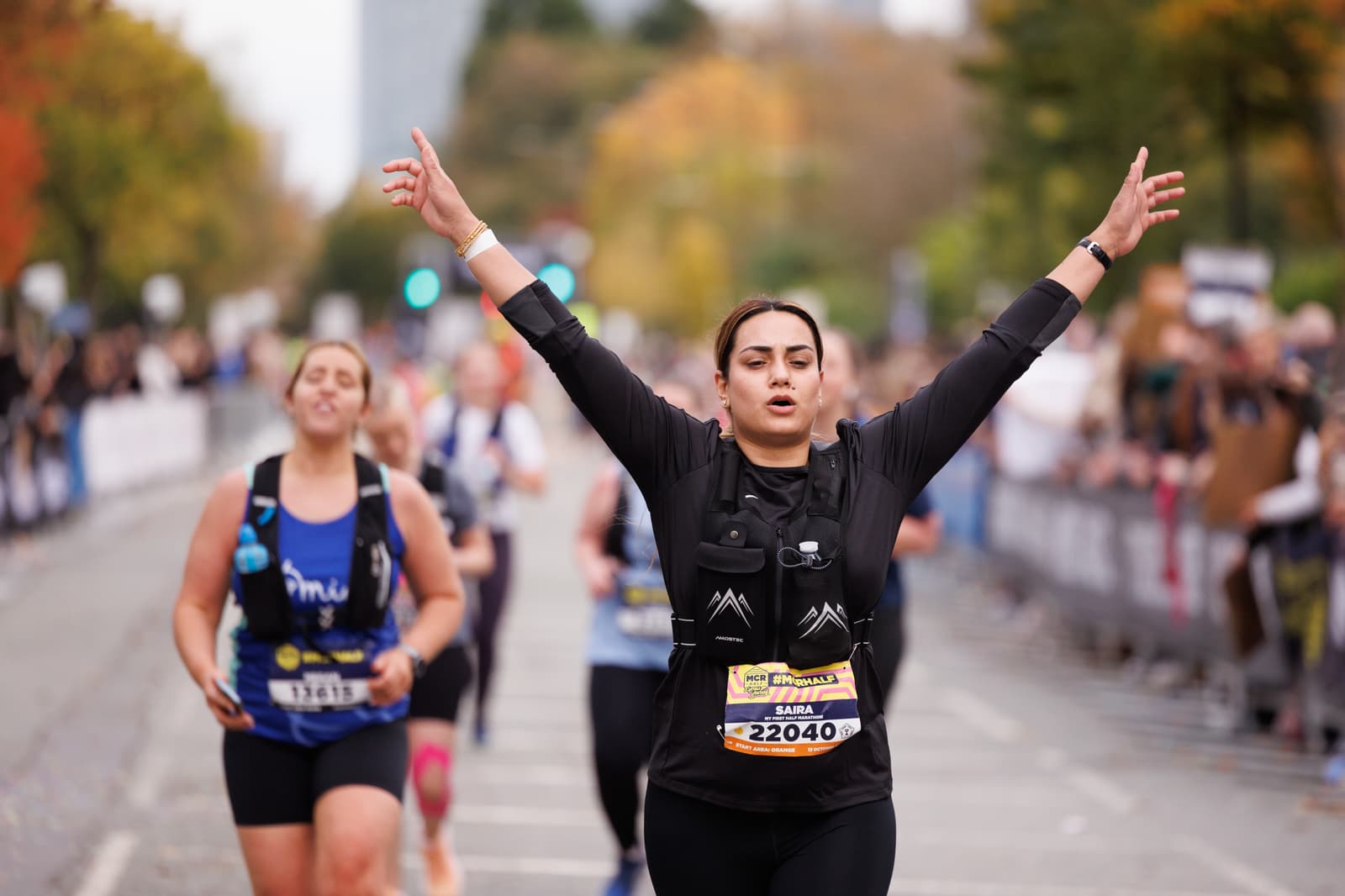 A runner crossing the marathon finish line with arms raised in triumph. She wears a bib number and looks elated. Other participants are seen running on the road. Trees with autumn foliage line the background.
