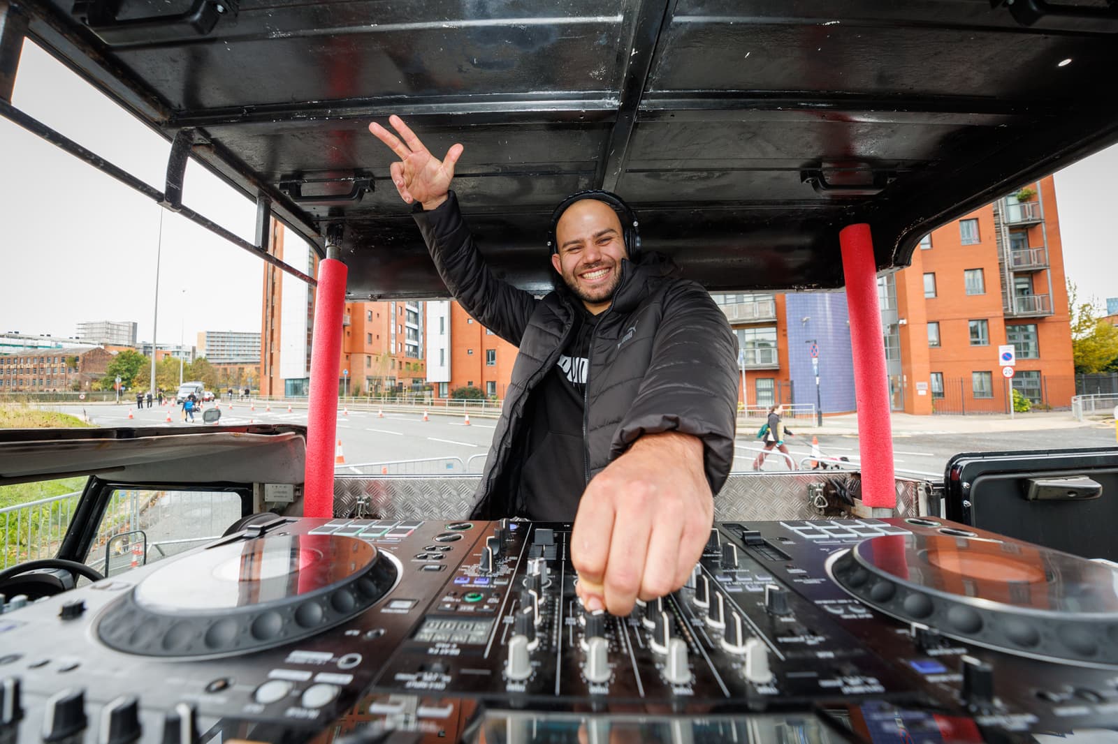 A DJ wearing headphones stands behind a large DJ mixer setup outdoors, smiling and gesturing with one hand. Buildings and a road are visible in the background. The scene reflects a lively, urban environment.