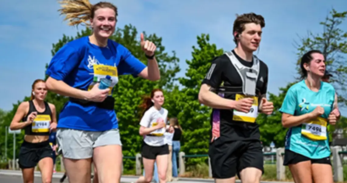 A group of runners participates in a race on a sunny day. A woman in a blue shirt smiles and gives a thumbs-up, while other runners, wearing various athletic gear, move alongside her. Trees and a clear blue sky are visible in the background.