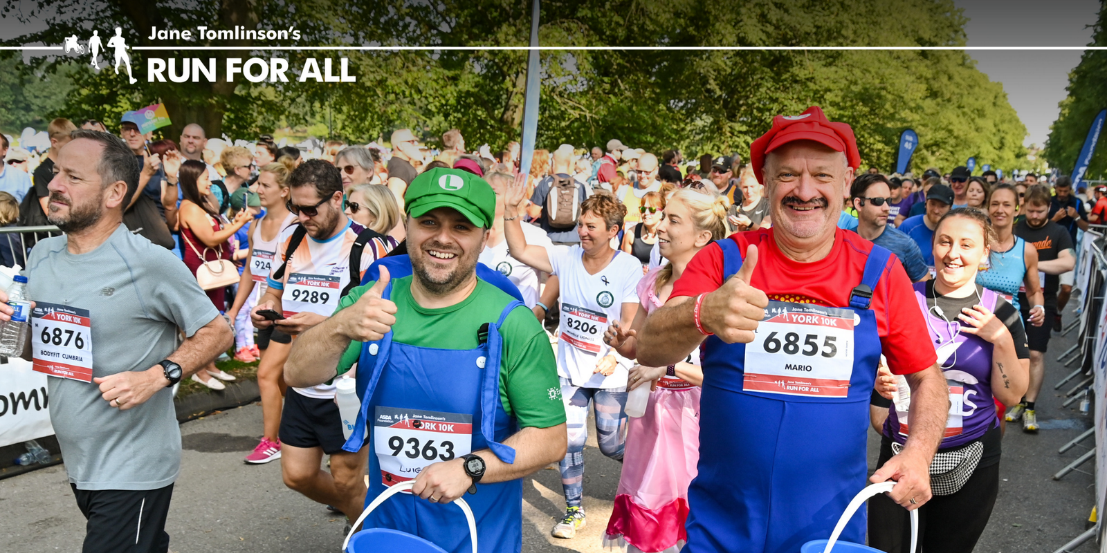 Participants are running in a "Run For All" event. Two men are in the foreground dressed as Mario and Luigi from "Super Mario." Both are smiling and giving thumbs up. Other runners, some in costumes, are visible in the background.