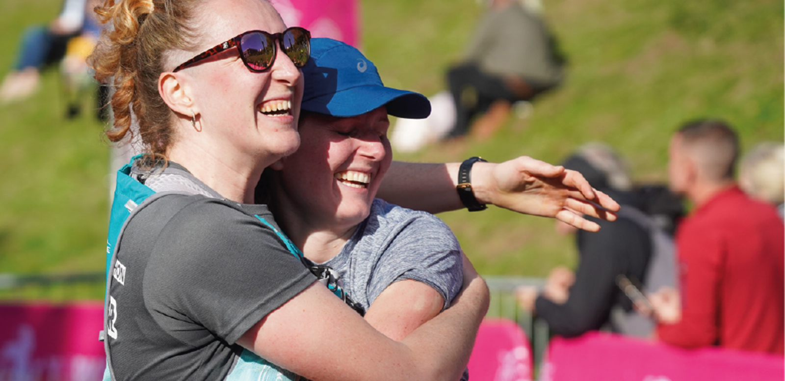 Two women share a joyful hug at an outdoor event. One is wearing sunglasses and a gray shirt with a bib number, while the other has a blue cap and a wristwatch. People and green grass are blurred in the background, suggesting a fun, active setting.