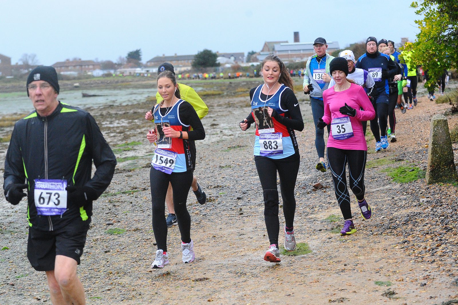 A group of runners, wearing numbered bibs, participate in an outdoor race on a path alongside a body of water. The runners are dressed in various athletic attire. In the foreground, a man in a black and neon-green jacket leads, with several others following closely behind.