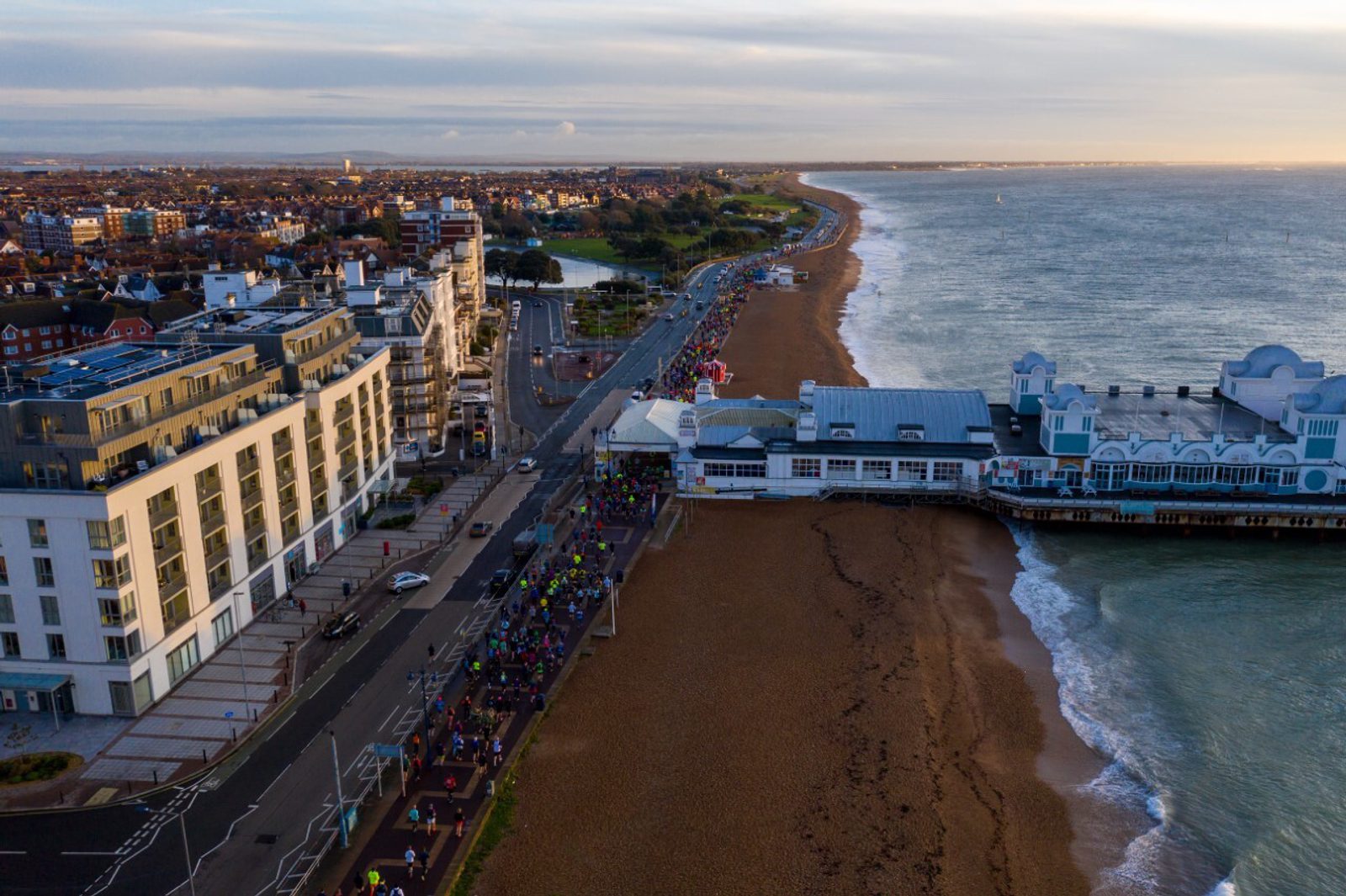 Aerial view of a seaside town with a long pier extending over the water. A crowd of people are gathered along the beach and adjacent road. Buildings line the waterfront, and waves gently wash onto the sandy shore under a partly cloudy sky.