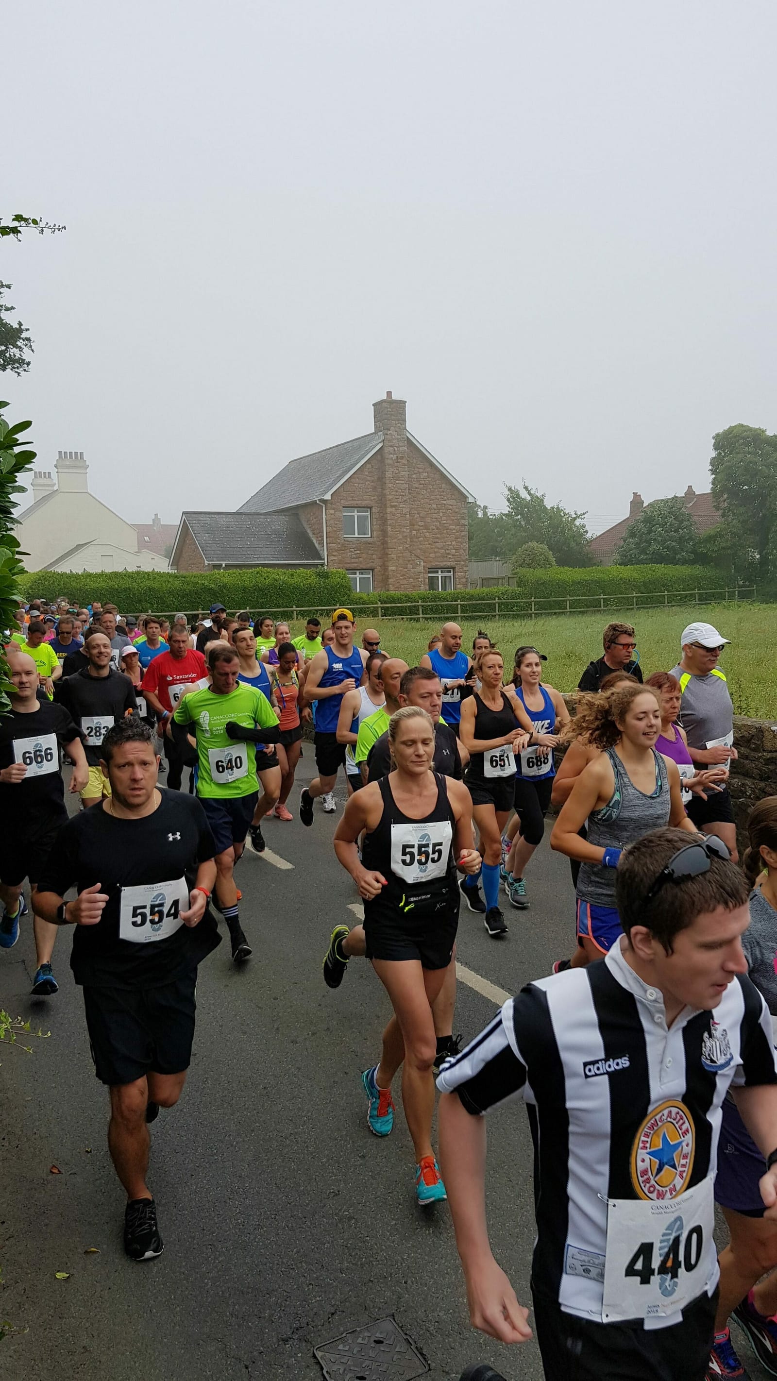 A large group of runners participating in a race on a foggy day, wearing numbered bibs. The event takes place on a road with houses and greenery in the background. The participants are dressed in various athletic outfits and appear focused on the run.