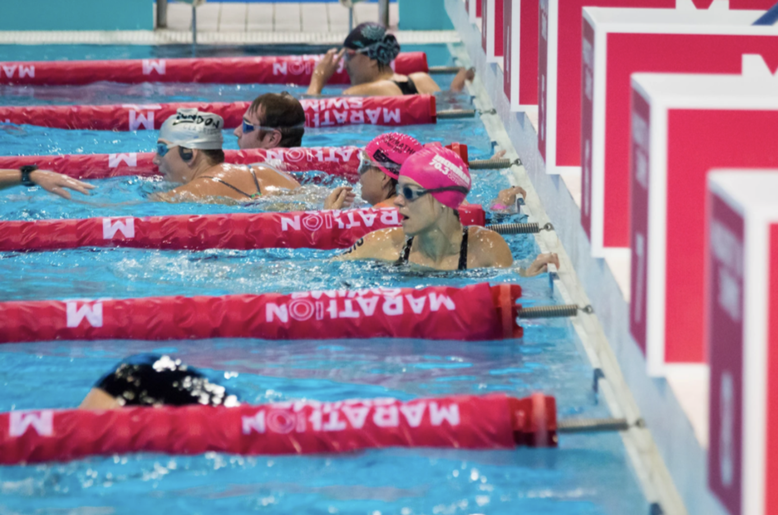 Swimmers wearing caps and goggles rest against the pool edge after finishing a race at a swimming competition, with red lane dividers marked "MARATON". The scene shows multiple lanes with competitors.