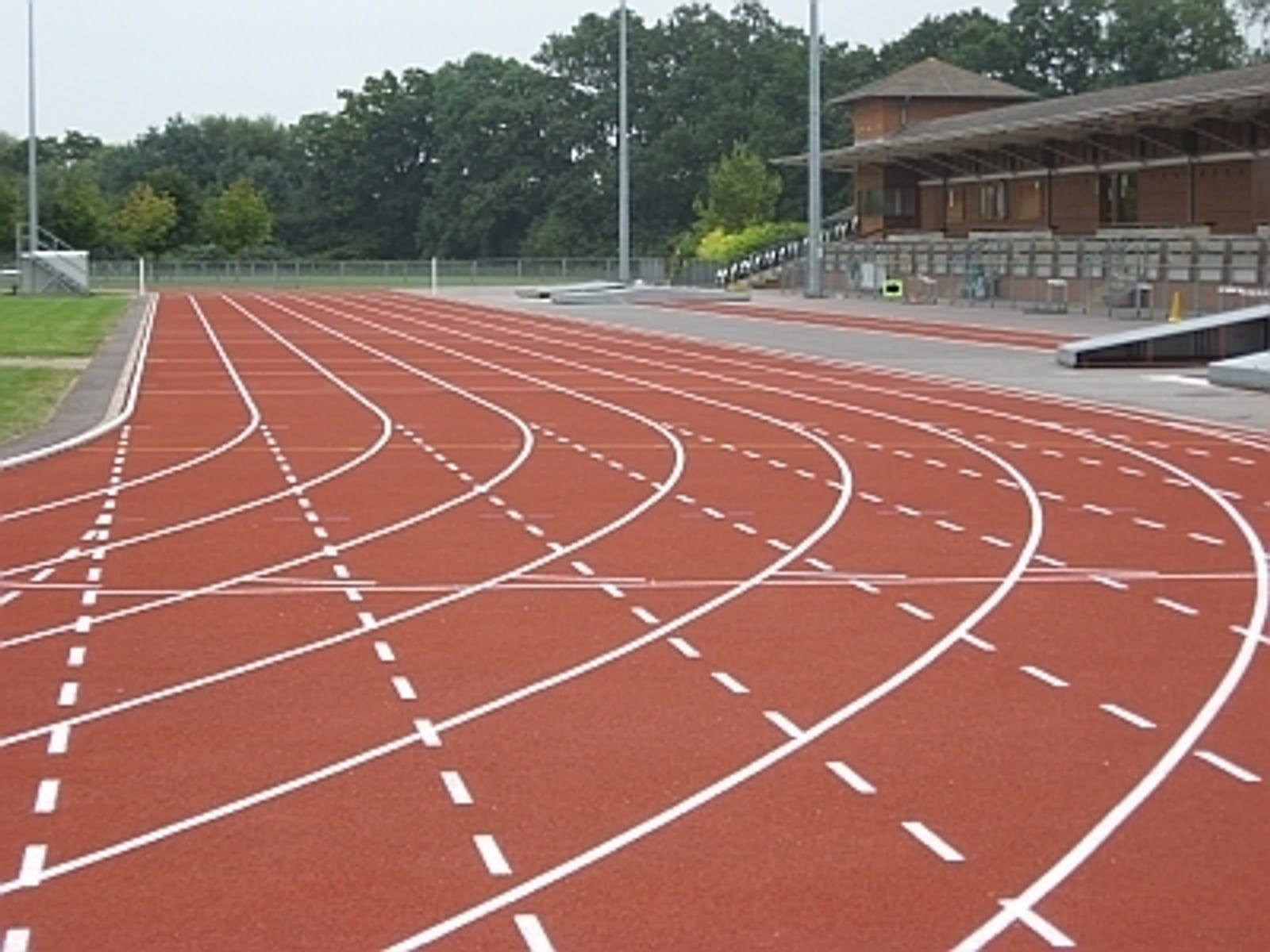An empty outdoor running track with multiple lanes, marked by white dashed lines, curves gently to the left. Adjacent to the track is a covered spectator stand with seats. Green trees and a grassy area line the background under an overcast sky.