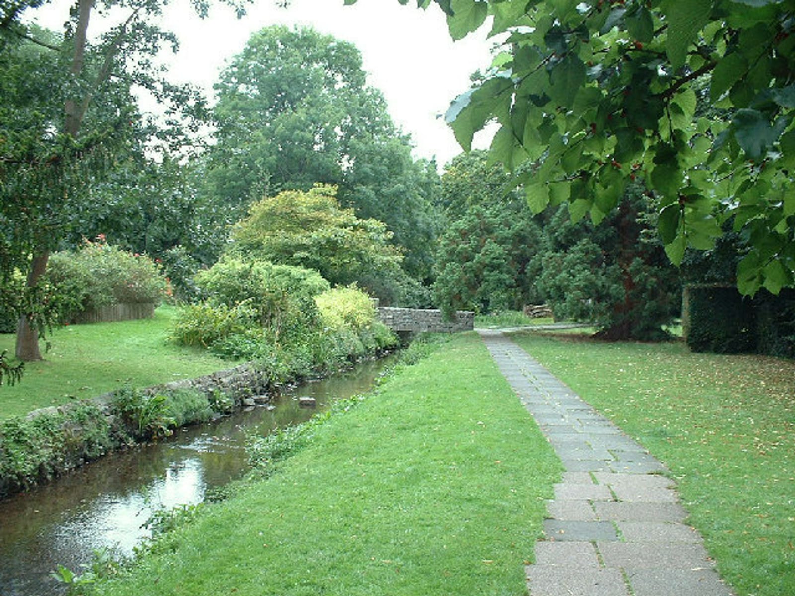 A serene park scene featuring a narrow stone pathway alongside a small, clear stream. The area is lush with green grass, various trees, and bushes. A stone bridge is visible in the distance, surrounded by the dense foliage of the park.