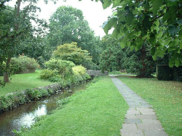 A serene park scene featuring a narrow stone pathway alongside a small, clear stream. The area is lush with green grass, various trees, and bushes. A stone bridge is visible in the distance, surrounded by the dense foliage of the park.
