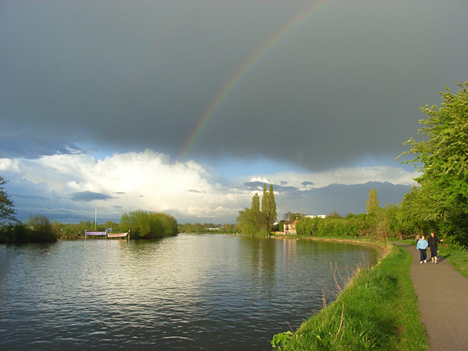 A serene riverside scene with a dark, cloudy sky partly giving way to sunlight, casting a rainbow. Two people walk on a path alongside the river, lined with green trees and bushes. A boat is moored on the opposite bank, and the atmosphere is calm.