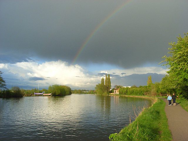 A serene riverside scene with a dark, cloudy sky partly giving way to sunlight, casting a rainbow. Two people walk on a path alongside the river, lined with green trees and bushes. A boat is moored on the opposite bank, and the atmosphere is calm.