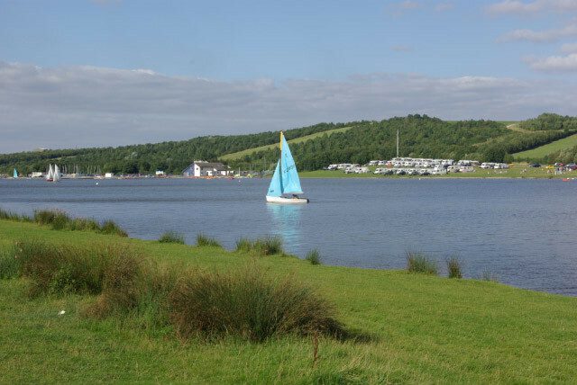 A serene lake with a single sailboat sporting a blue sail gliding on the water. The background features rolling green hills, a cluster of white buildings, and scattered trees under a blue sky with scattered clouds.