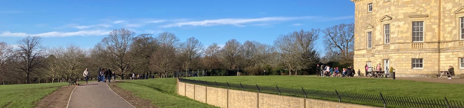 Pathway leading to a large stone building on the right, surrounded by a lawn. People are walking and sitting outside, with trees in the background and a clear blue sky above.