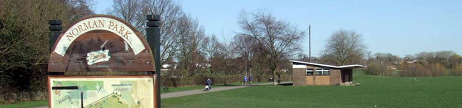A park on a clear day featuring a green lawn, trees without leaves, and a path along which a few people are walking. There is a brown and white signpost that says "Norman Park" and a small building can be seen in the background.