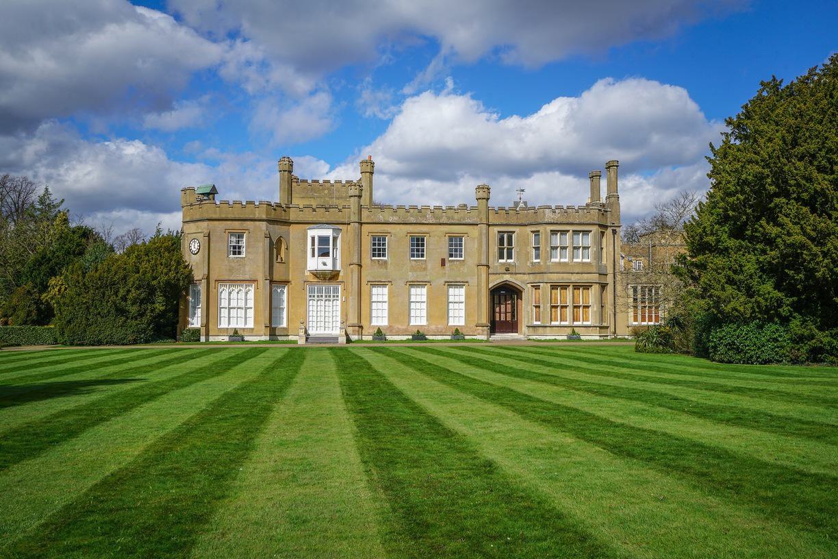 A historic mansion with beige stone walls, symmetrical architecture, and turrets. The manicured lawn in the foreground is striped with alternating light and dark green grass. The sky is blue with scattered clouds, and trees surround the building.