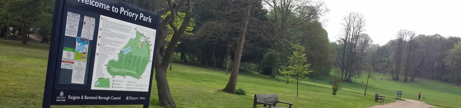 A large sign at the entrance of Priory Park displays a detailed map and information. There is a wooden bench nearby, and a path leading through green, well-maintained grassy areas with scattered trees. The sky is clear and the park appears peaceful and inviting.