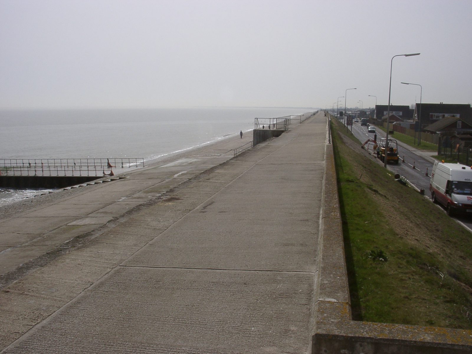 A long concrete seawall stretches alongside a foggy shoreline with calm seas on the left and a road bordered by grassy areas and buildings on the right. A few vehicles and people are visible in the distance. The sky is overcast, giving a greyish hue to the scene.