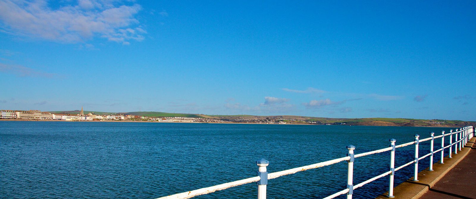 A scenic view of a coastal town from a pier under a clear blue sky. The calm sea stretches out towards the horizon, where low hills and the town's buildings are visible. A white handrail runs along the edge of the pier in the foreground.
