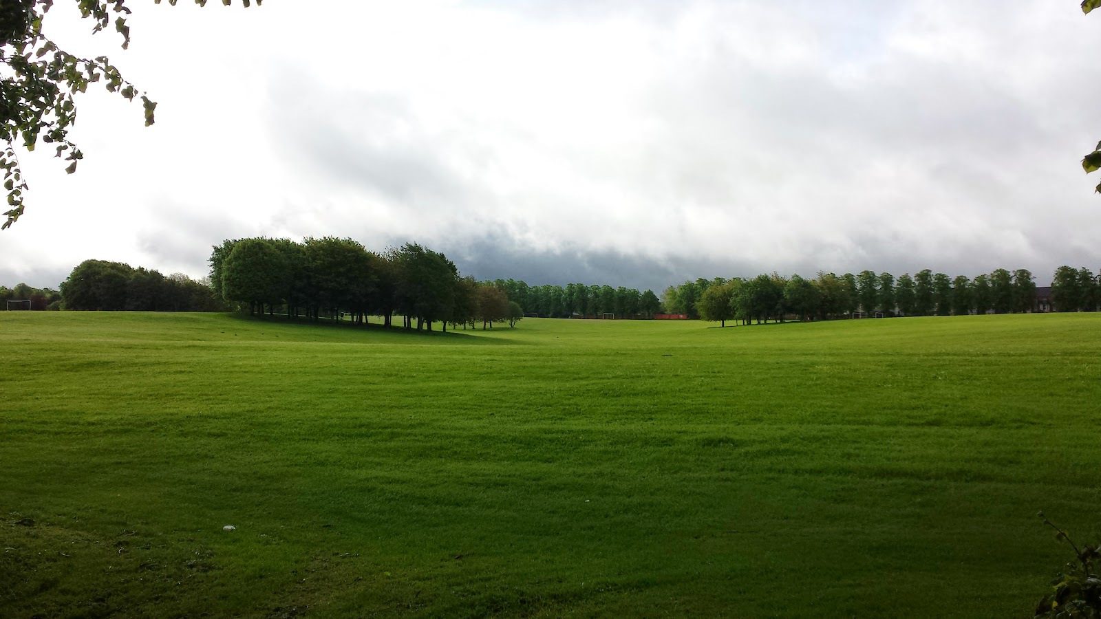A vast green field stretches into the distance, bordered by trees. The sky above is partly cloudy, with patches of blue sky visible through the clouds. The grass is lush and well-maintained, creating a serene and open landscape.