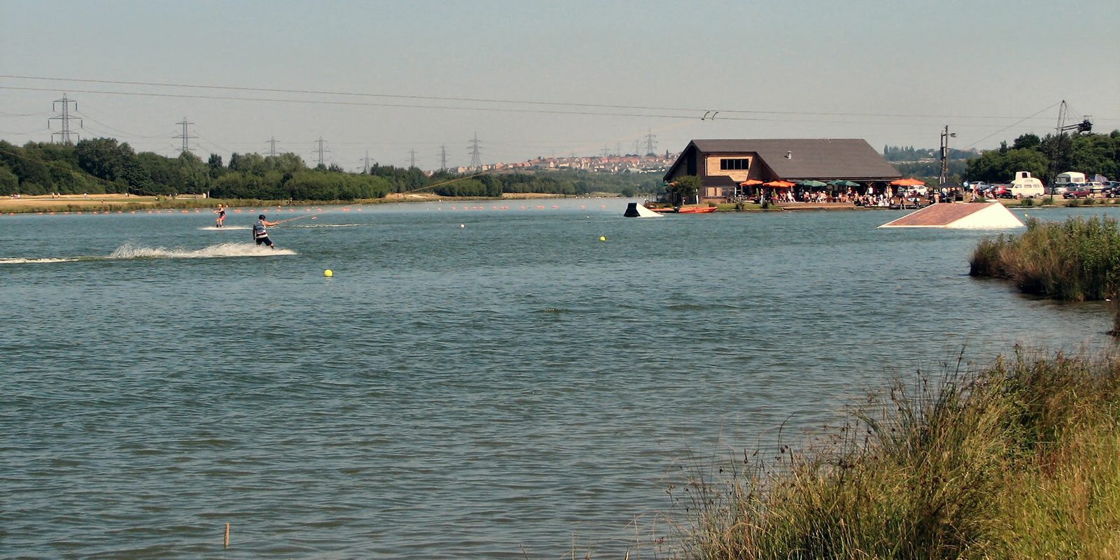 A view of calm water bordered by grass, with a person wakeboarding in the distance. Further back, there is a wooden building on the shore with people gathered outside. In the background, there are power lines and rolling hills under a clear sky.