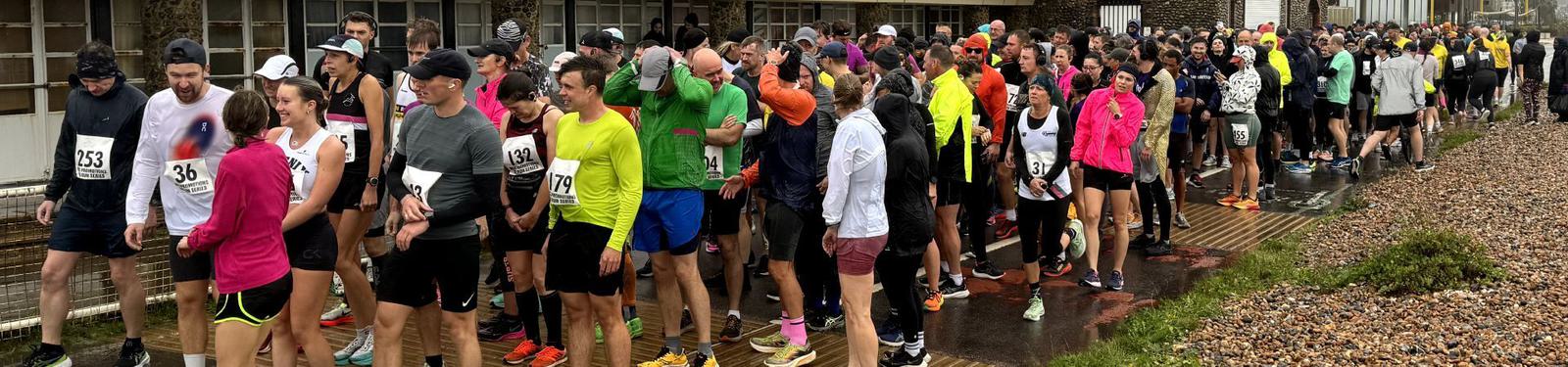 A large group of runners in colorful attire gather at the starting line of a race. Some are wearing numbers on their shirts. The path is wet, indicating recent rain, and the overcast sky adds to the damp atmosphere.